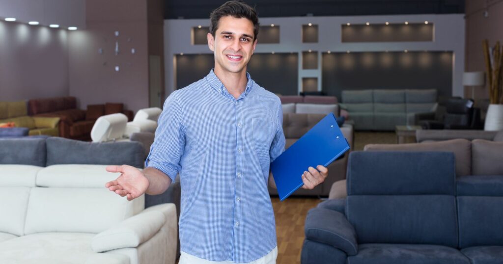 A man in a furniture store, holding a clipboard, appears to be a modern sofa seller.