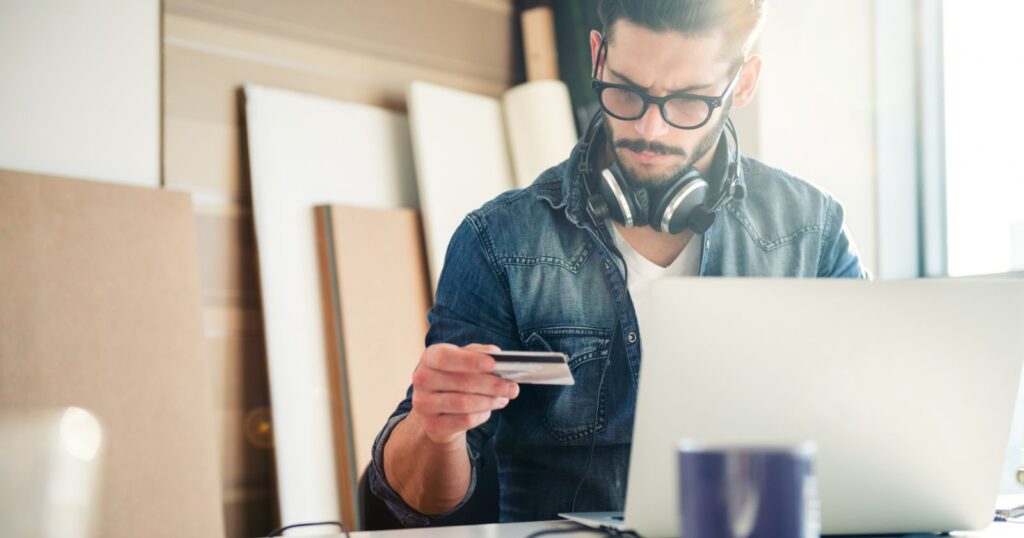 A man with headphones and glasses using a laptop and a credit card to explore Different Types of Checking Accounts.