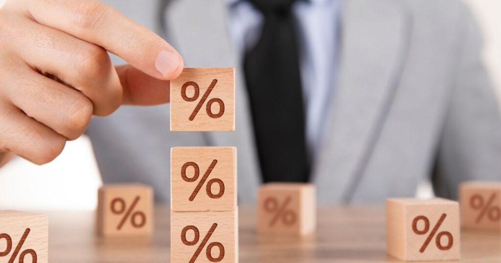 A man in a suit strategically arranges wooden blocks with percent signs, symbolizing factors affecting home loan interest rates.