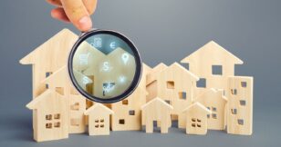 A hand holds a magnifying glass over wooden houses with icons, symbolizing the search for home loan rates in today's market.