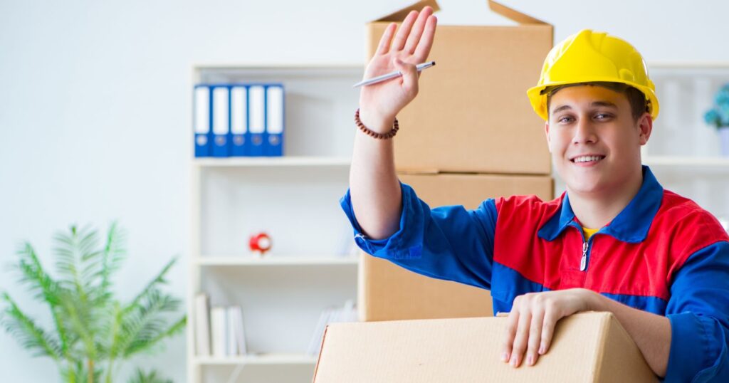 A man in a hard hat and red shirt holds a box. Image representing 'The Etiquette of Tipping Movers'.