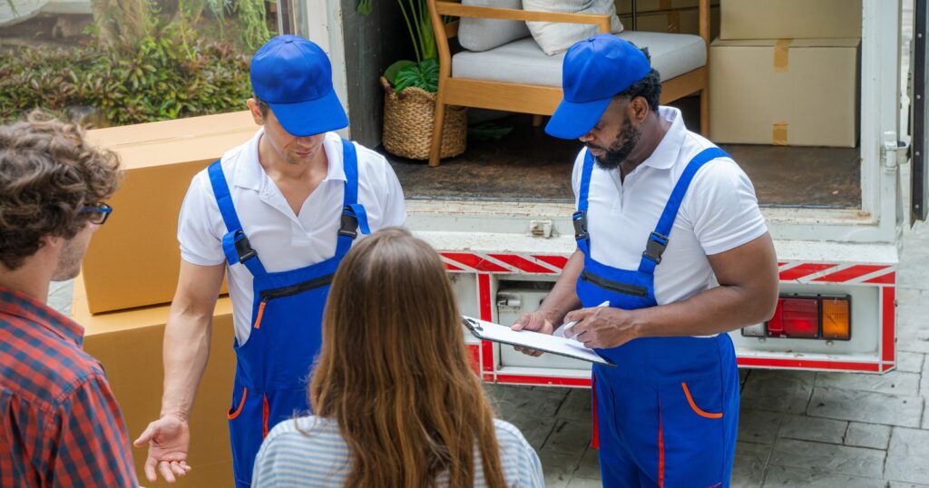 Three people in blue overalls standing near a moving truck. They appear to be discussing "The Impact of Your Tip."