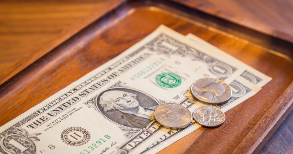 A wooden tray with $3 and 2 coins, representing suggested tipping amounts on a table.